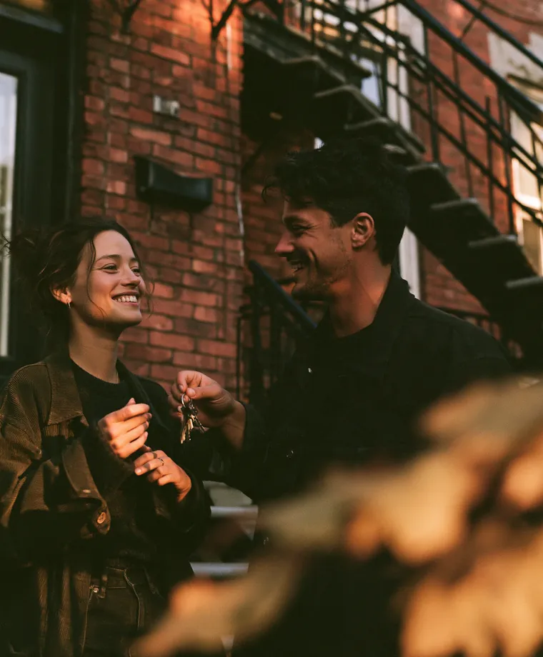 Jeune couple souriant qui tient les clés de son premier logement devant un immeuble en brique